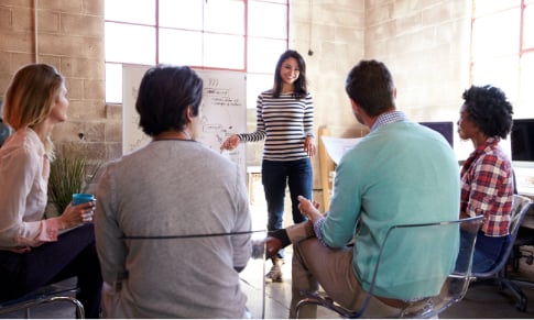 Presenter standing in front of an audience presenting a chart on a whiteboard.